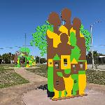 One part of a three-piece flat metal sculpture. This section has houses, trees and silhouettes that are yellow, brown and green.